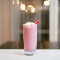 A glass of strawberry milkshake with whipped cream and a slice of strawberry on top placed on a white surface