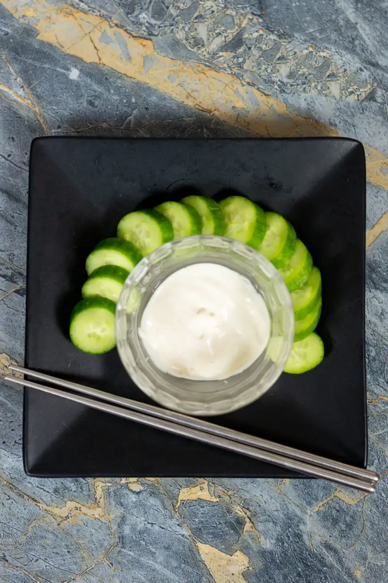 Japanese white sauce in a clear bowl surrounded by cucumbers on a black plate with chopsticks on it