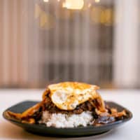 A chopped steak over white rice topped with a fried egg placed on a black plate