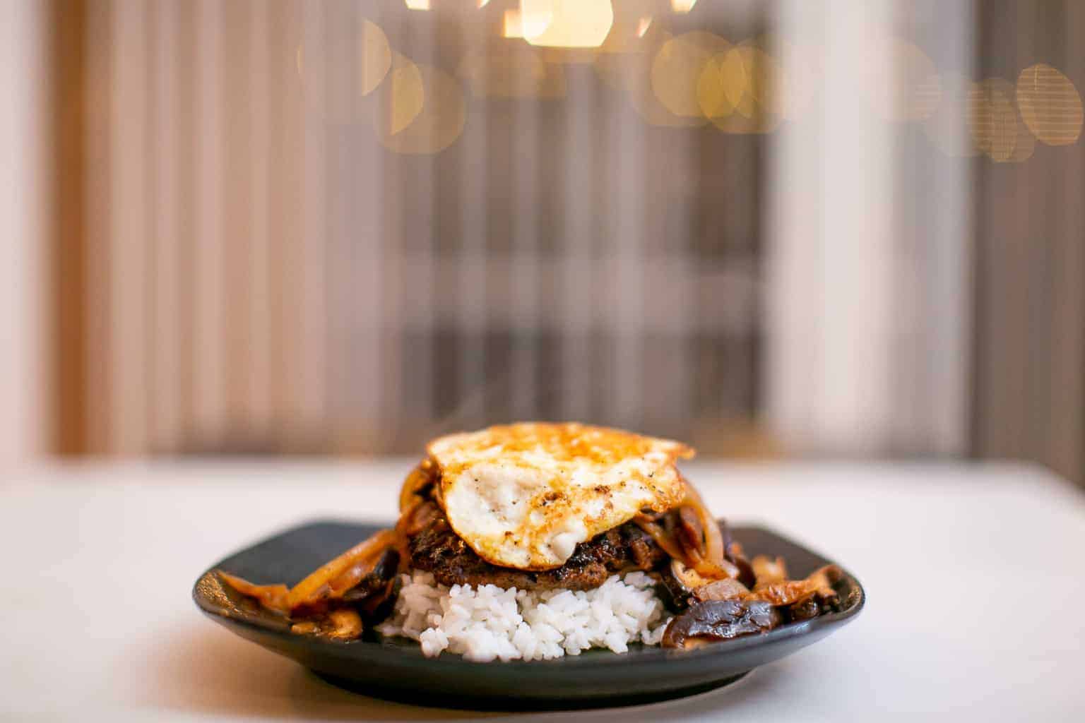 A chopped steak over white rice topped with a fried egg placed on a black plate