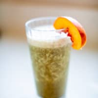 Blended green smoothie in clear glass with a slice of peach placed on a white surface