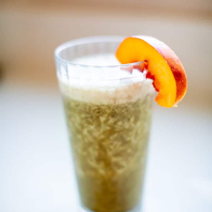 Blended green smoothie in clear glass with a slice of peach placed on a white surface