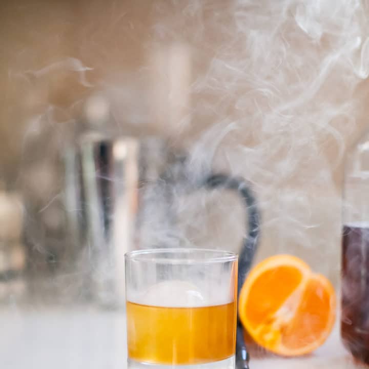 A glass of maple bourbon smash with smoke near a slice of orange fruit placed on a white surface