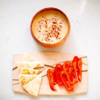 Roasted garlic hummus in a brown wooden bowl near a pita bread and red bell pepper placed on a charcuterie board