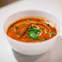 Rustic tomato soup garnished with basil leaves served in a white bowl placed on a white surface
