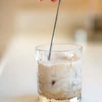 A person stirring a glass of white Russian cocktail using a silver stirrer