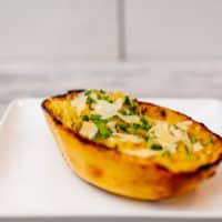 Spaghetti squash garnished with cheese and herbs on a square white plate placed on a white wooden table