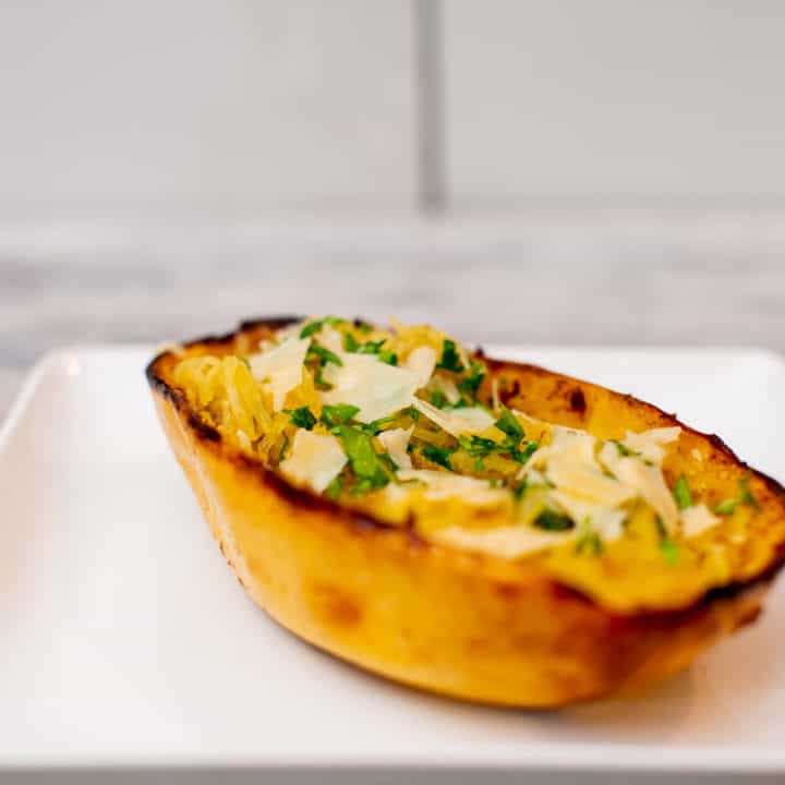 Spaghetti squash garnished with cheese and herbs on a square white plate placed on a white wooden table