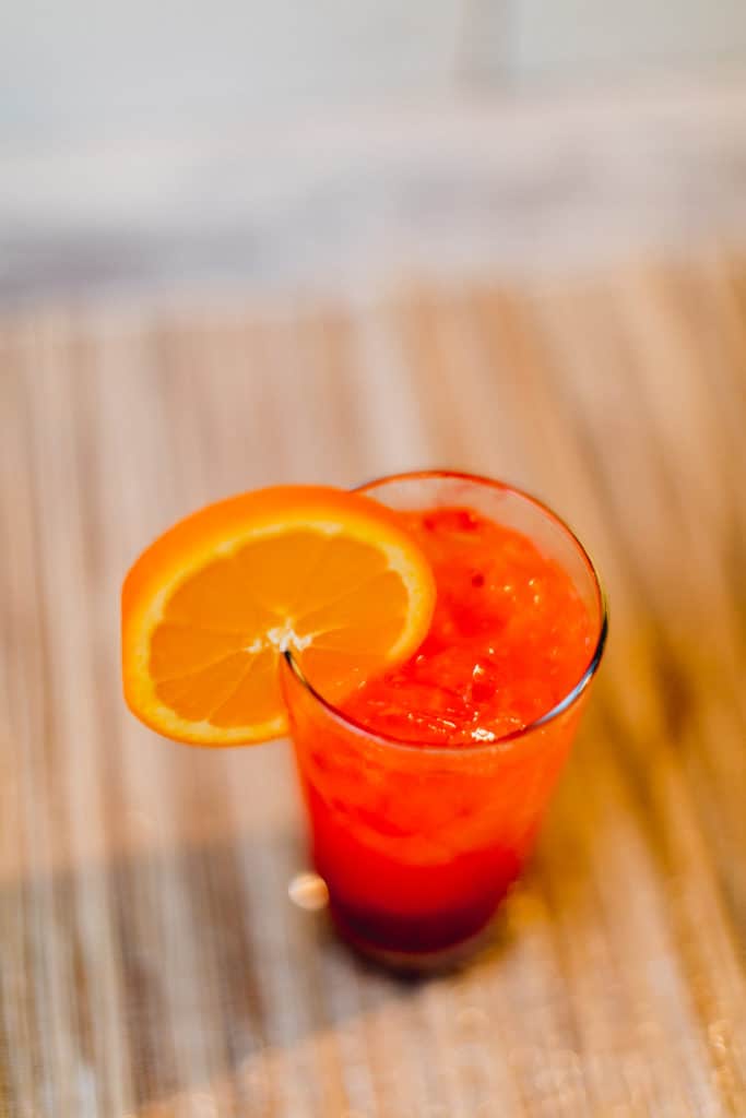 A top view of a tequila sunrise cocktail with a slice of orange in a glass placed on a brown surface