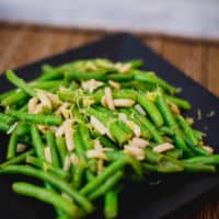 A close-up of a green bean salad topped with nuts served on a black plate placed on a brown placemat