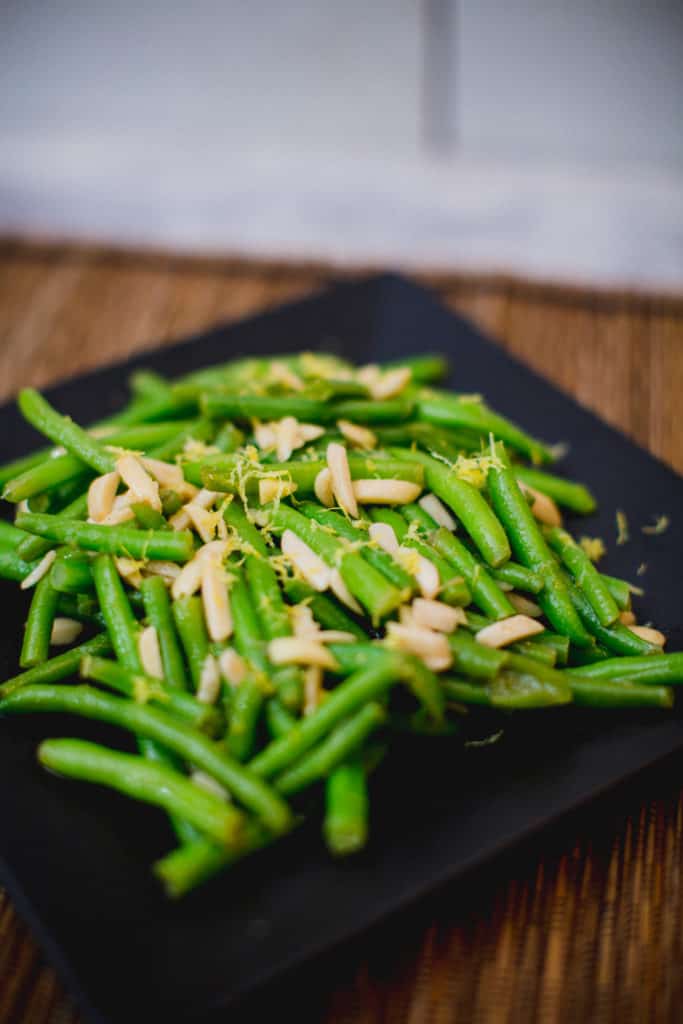 A close-up of a green bean salad topped with nuts served on a black plate placed on a brown placemat