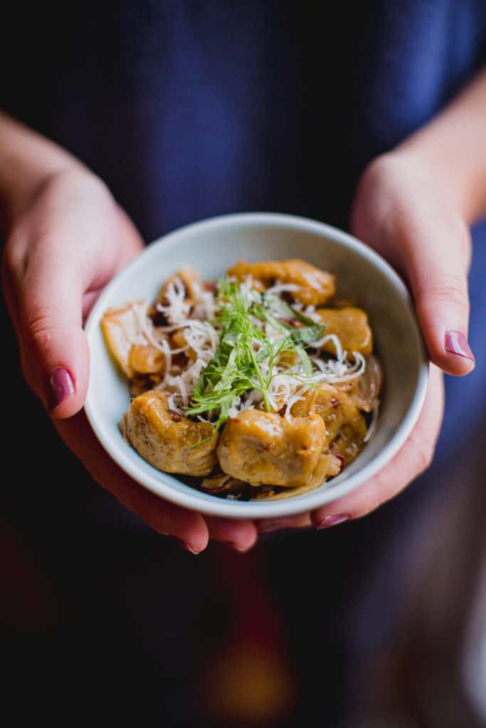 A person holding a bowl of butternut squash gnocchi garnished with parmesan cheese and herbs