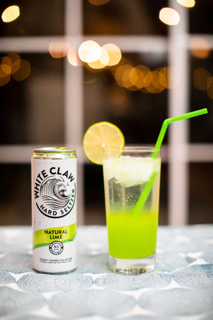 A can of White Claw seltzer placed beside a glass of lime White Claw cocktail that has a straw and a slice of lemon on its glass