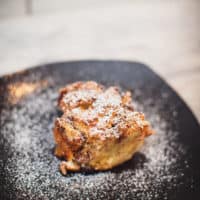 A slice of eggnog bread pudding is served on a square black plate put on a wooden surface