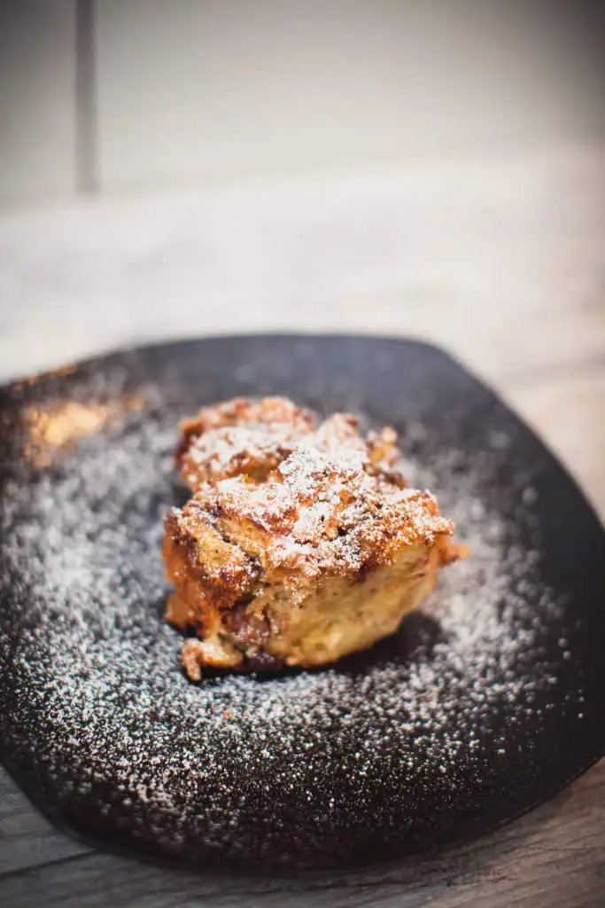 A slice of eggnog bread pudding is served on a square black plate put on a wooden surface