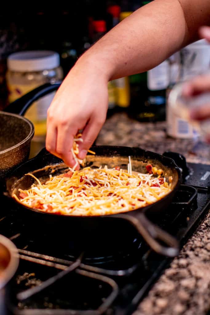 Person adding cheese and bacon while preparing homemade creamed corn in a cast iron pan