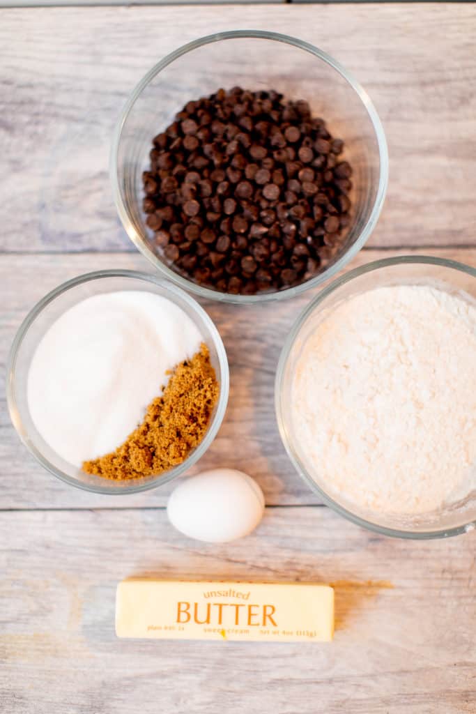 A bowl of flour, chocolate chips, egg, butter, and white and brown sugar placed on a table