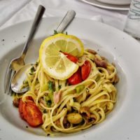 Lemon and capers pasta sauce garnished with lemon and herbs on a white plate with a spoon and fork placed on a white tablecloth