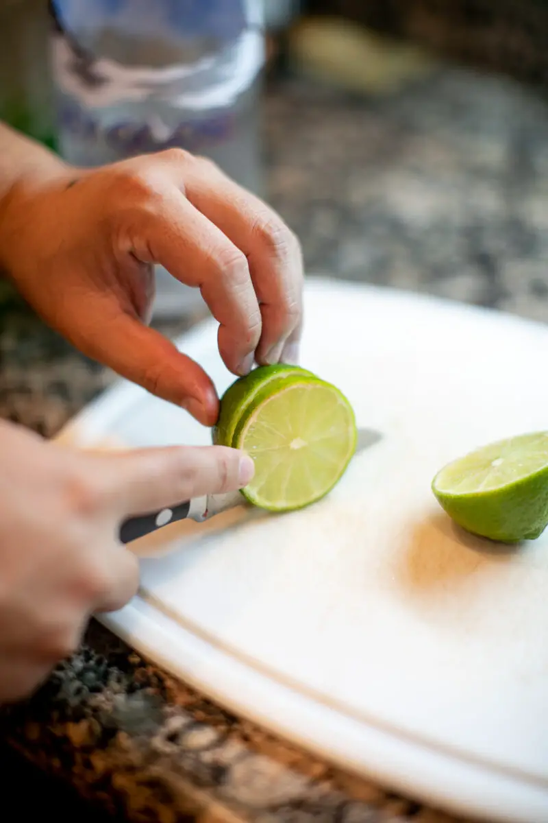 Man using a knife to cut a lime into round slices on a white cutting board in the kitchen