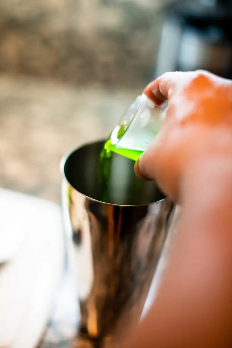 Man pouring Midori into a shaker for a mixed drink on the kitchen table