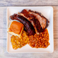 A top view of a sous vide baby back ribs with beans and dinner rolls served on a white plate put on a wooden surface