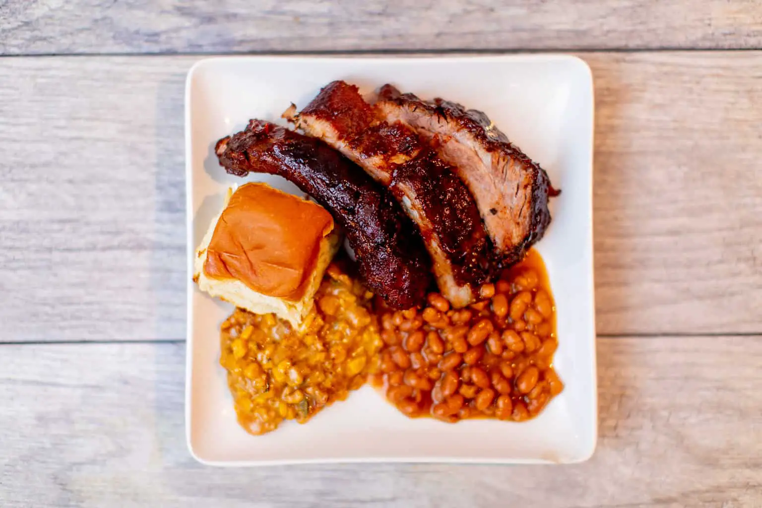 A top view of a sous vide baby back ribs with beans and dinner rolls served on a white plate put on a wooden surface