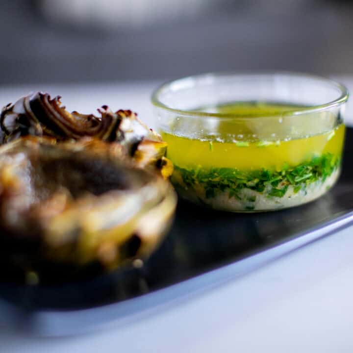 Roasted artichokes on a black plate with lemon garlic butter sauce in a small glass bowl placed on a white table