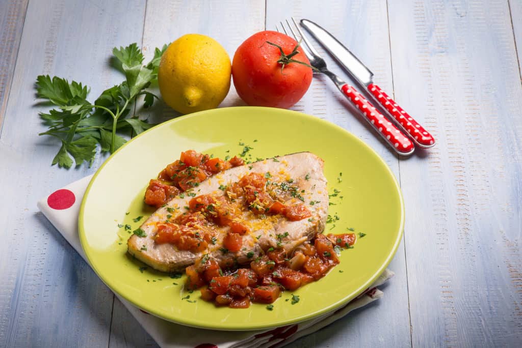 Lemon garlic swordfish garnished with tomatoes served on a yellow plate beside a fresh lemon and tomato placed on a polka dot cloth