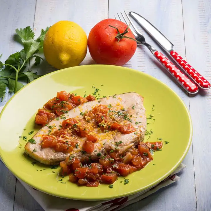 Lemon garlic swordfish garnished with tomatoes served on a yellow plate beside a fresh lemon and tomato placed on a polka dot cloth
