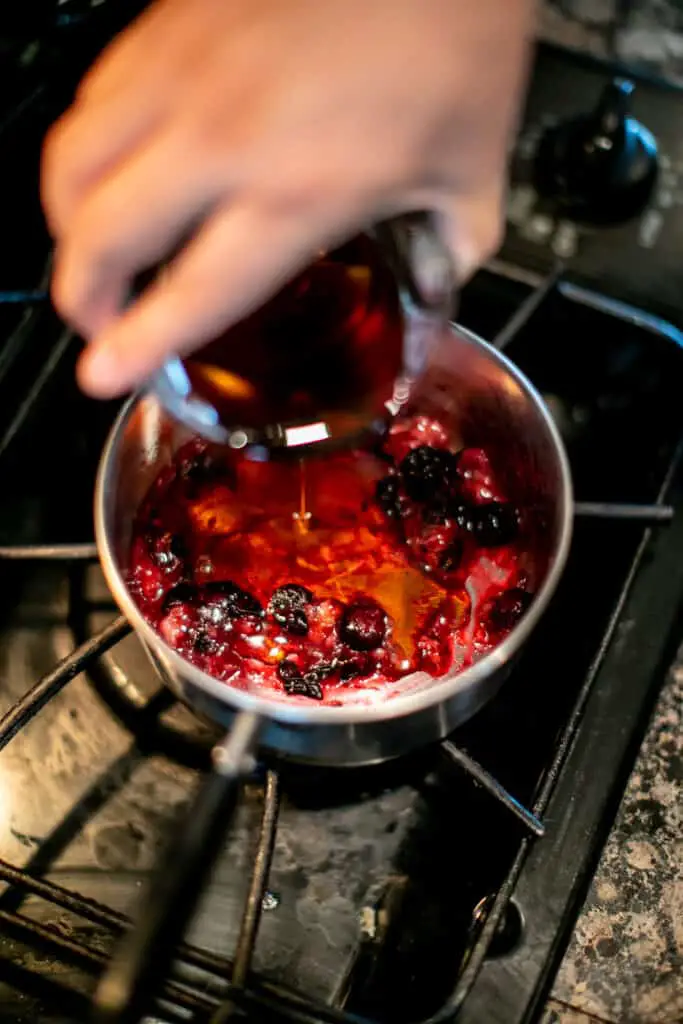 Maple syrup being poured in blueberry mixture while being cooked in a saucepan