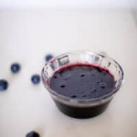Blueberry syrup in a small clear bowl near blueberries placed on a white surface