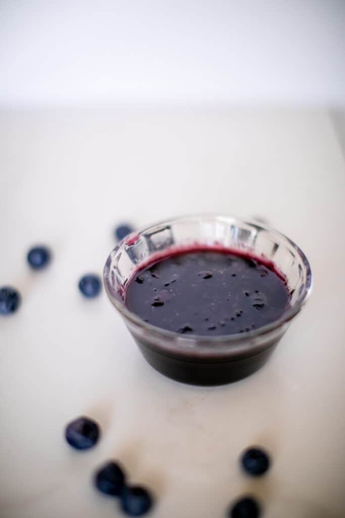 Blueberry syrup in a small clear bowl near blueberries placed on a white surface