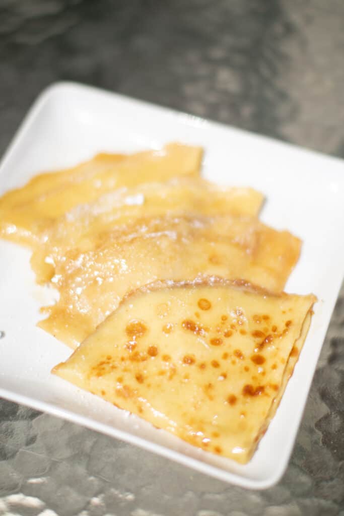 Crepes suzette topped with powdered sugar served on a white plate placed on a glass table