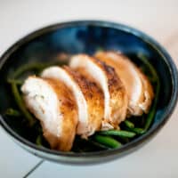 Oven-roasted whole chicken in a bed of vegetables served in a black bowl placed on a white surface