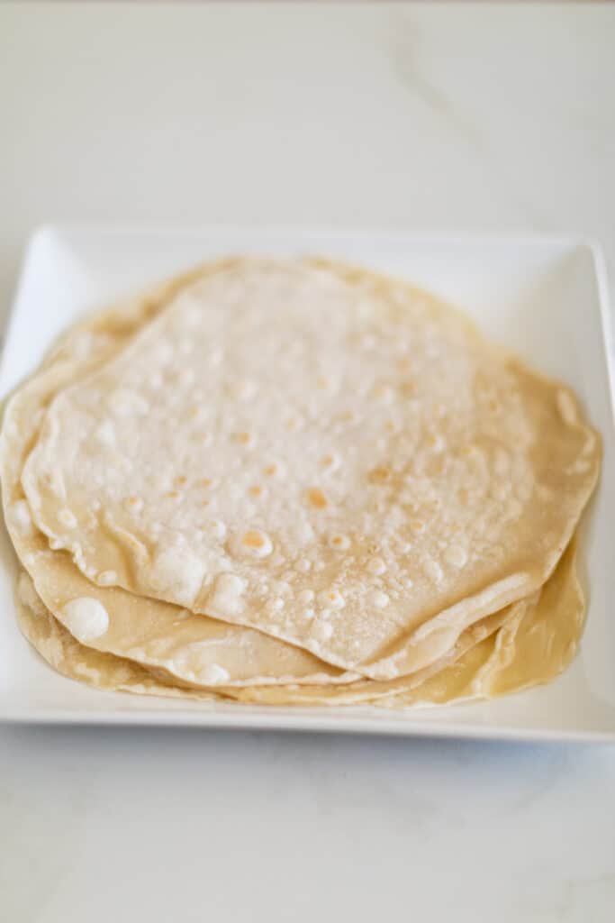 A pile of soft flour tortillas served on a square white plate placed on a white surface