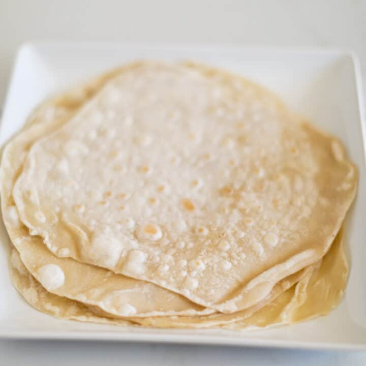A pile of soft flour tortillas served on a square white plate placed on a white surface