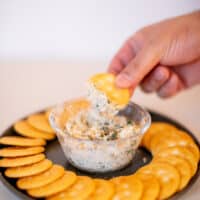 A person dipping a cracker on a spinach artichoke served in a small glass bowl