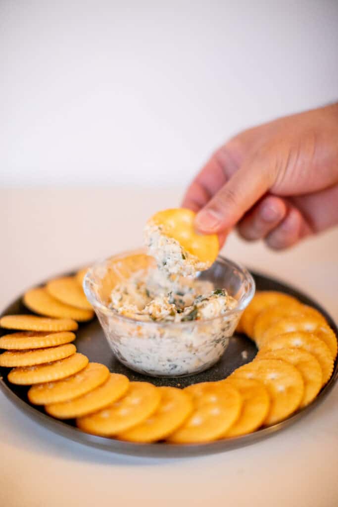 A person dipping a cracker on a spinach artichoke served in a small glass bowl