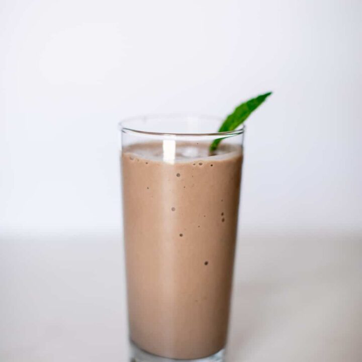 Chocolate peanut butter banana smoothie with mint leaf served in a clear tall glass placed on a white surface