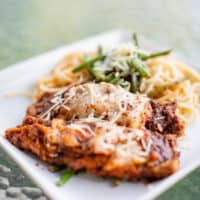 Breaded fried cutlet of veal topped with tomato sauce and mozzarella cheese served on a square white plate placed on a glass table