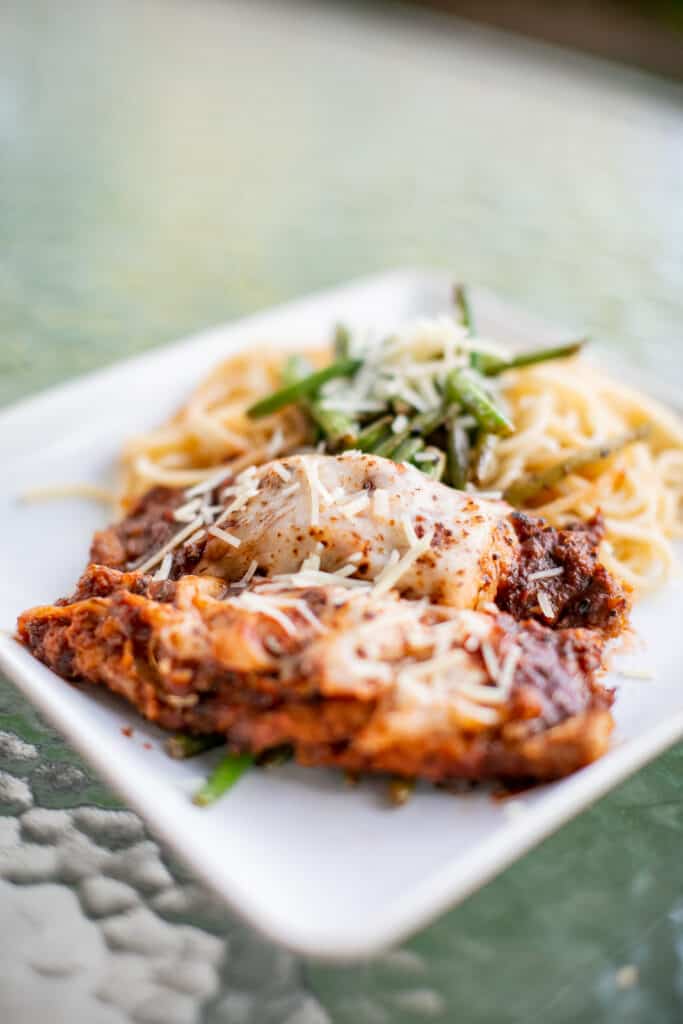 Breaded fried cutlet of veal topped with tomato sauce and mozzarella cheese served on a square white plate placed on a glass table