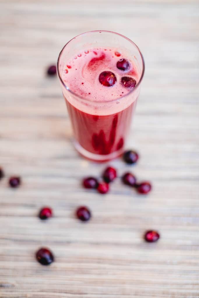 A top view of cranberry juice in a tall glass with fresh cranberries around a wooden surface