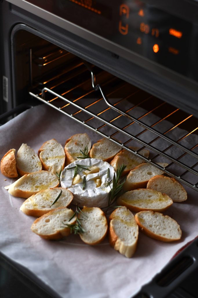 An open oven with goat cheese in the center of the pan with bits of bread