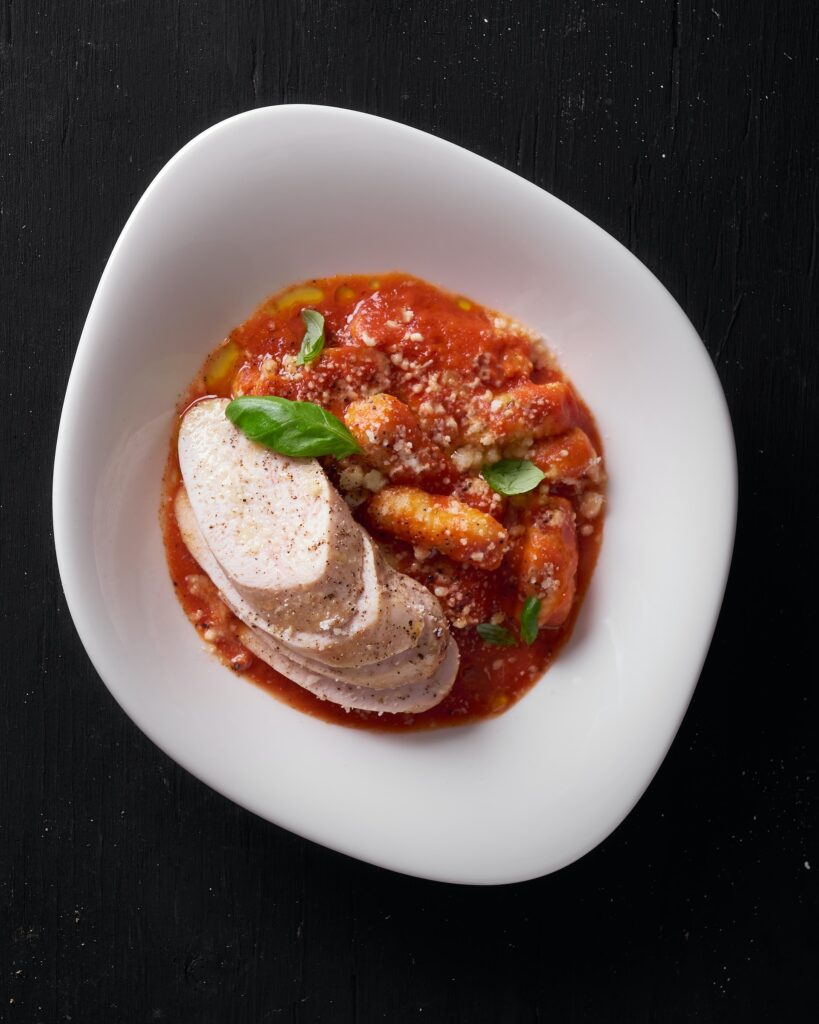 A top view of Gnocchi alla Sorrentina with Sous Vide Chicken on a white plate placed on a black wooden table