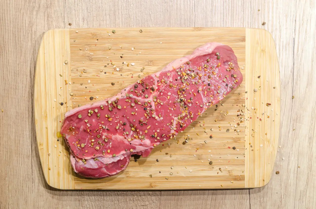 A slice of meat sprinkled with ground peppercorns on a brown wooden chopping board