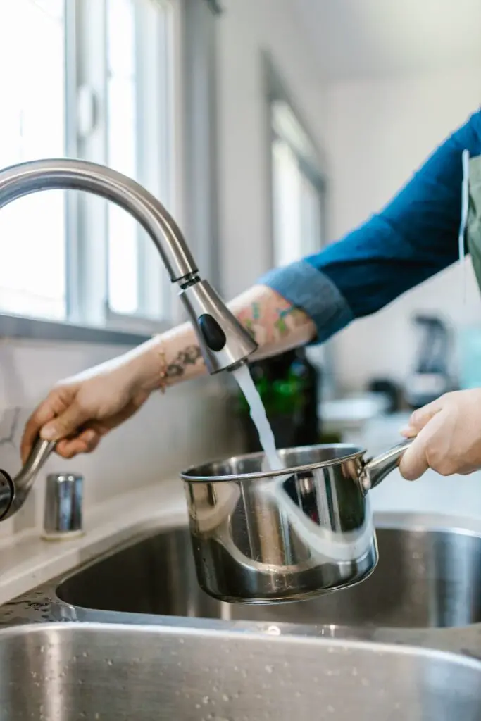A person in blue long sleeves filling a stainless pot with water in the silver faucet