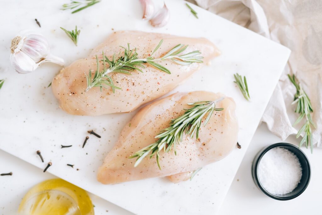 Two sliced raw chicken breasts with herbs and spices on a white ceramic plate