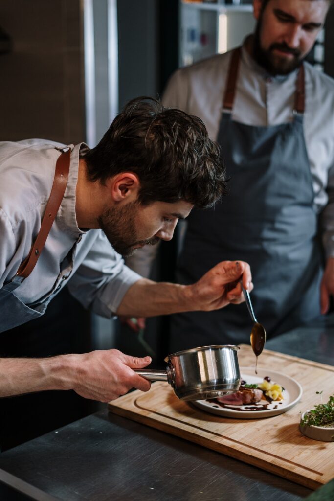 A man in long sleeves polo and a blue apron plating a dish on a white plate placed on a brown wooden board