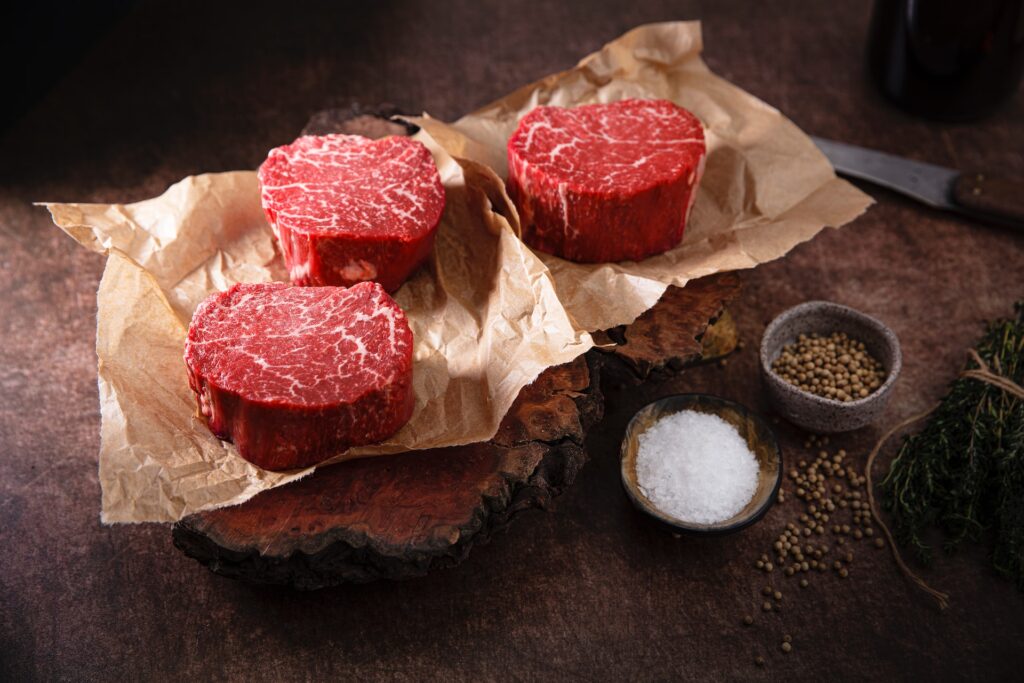 Three sliced steaks on brown parchment paper with different spices on the side placed on a brown wooden table