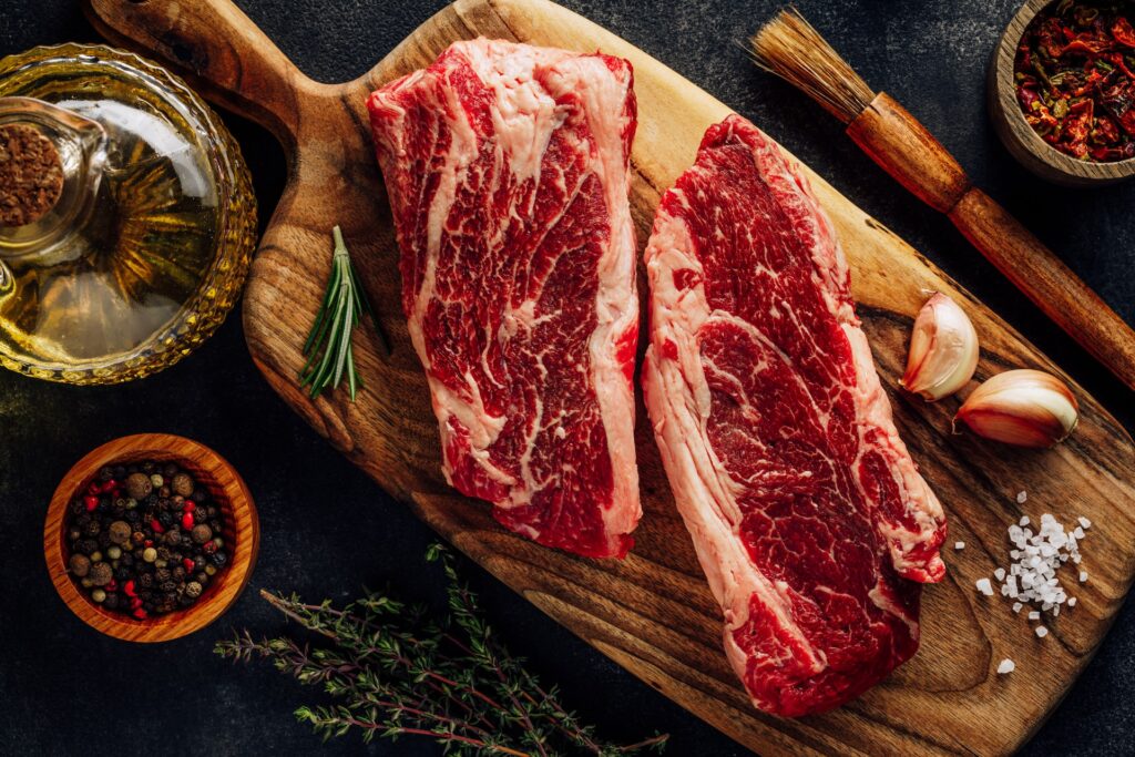 Top view of raw marbled steak with different spices and spices on a brown wooden cutting board placed on a black table